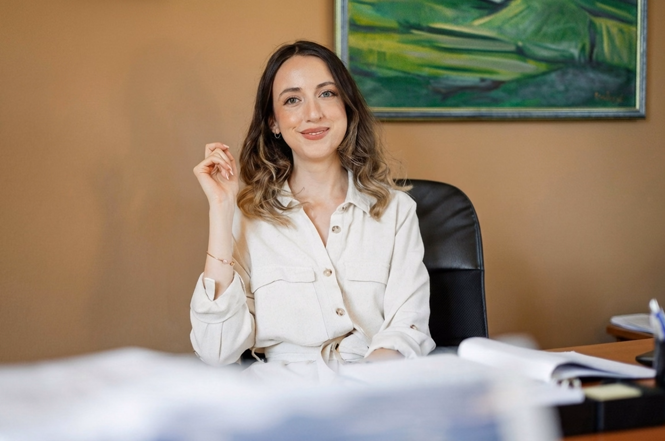 Smiling woman with wavy hair sitting on a black office chair in front of a desk with papers, with a green landscape painting on a beige wall behind her.